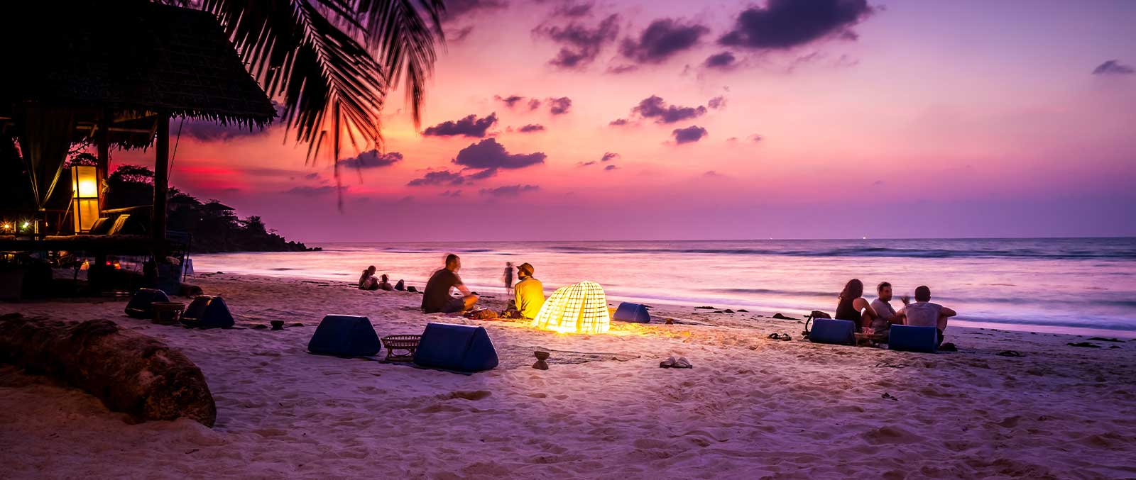 People are sitting at the bank of river in front of palm trees, at the time of sunset and lights up with lanterns