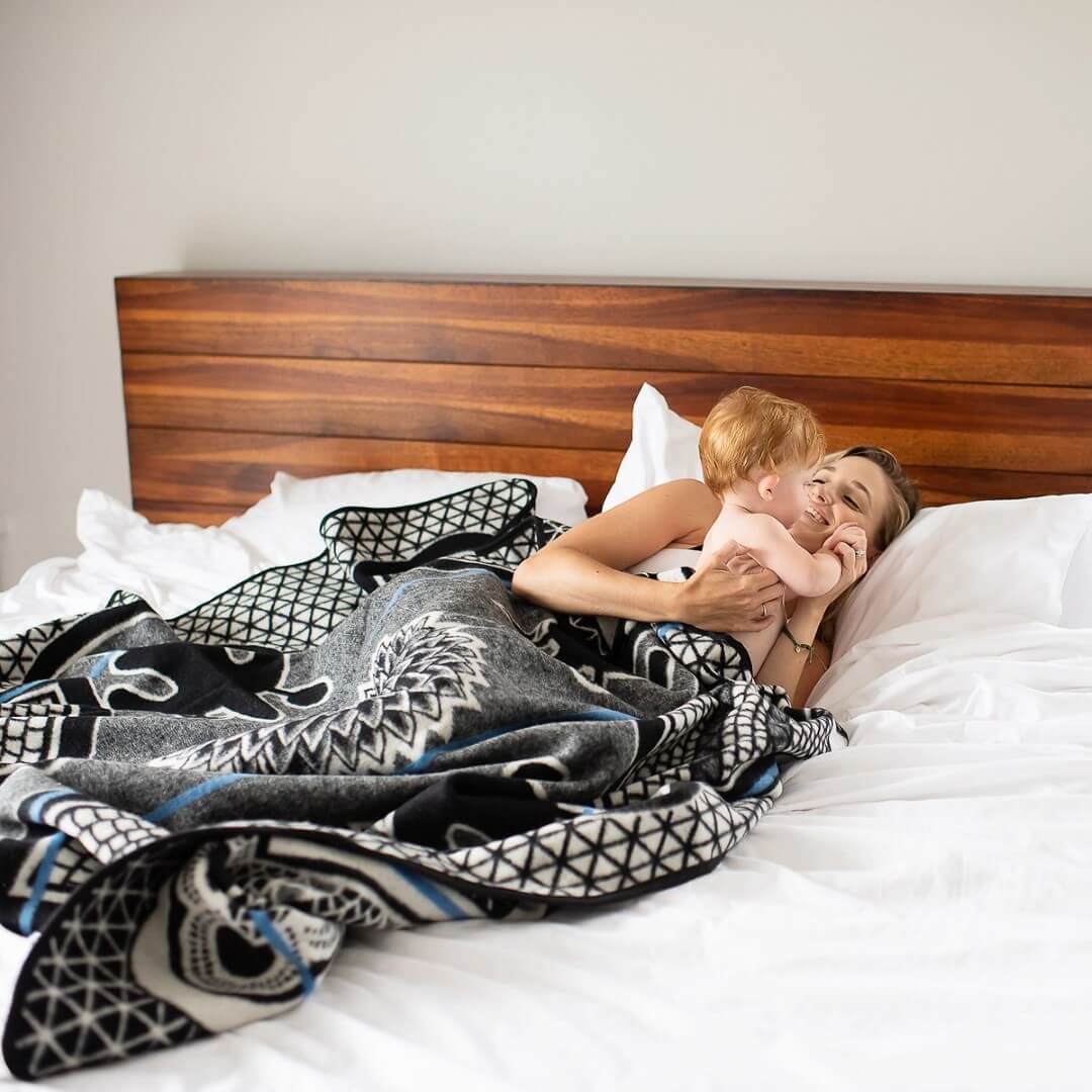 Women and child playfully cuddle on a big comfy bed lying under Basotho Heritage blanket Kharetsa blanket, wrap or throw sustainably and ethically made in South Africa in South Africa chromatic royal blue and cobalt throw draped across a  bed in modern home 