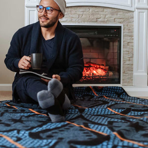 Man relaxing infront of fire reading a book with legs crossed sitting on an Authentic SeannaMarena blue and gold Basotho Heritage Wool Blanket and wrap or throw sustainably and ethically crafted in South Africa draped over modern house in South Africa