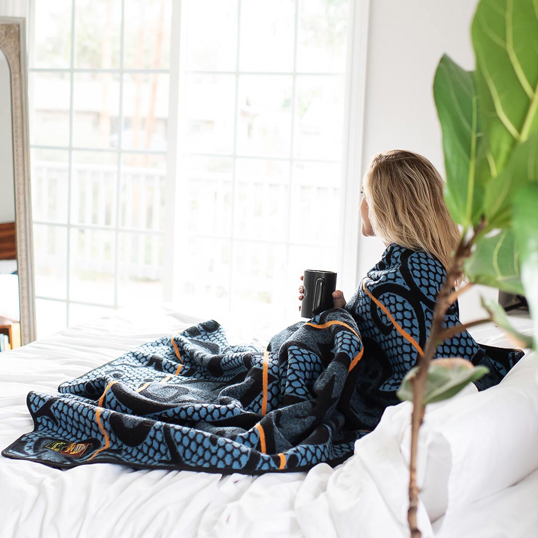 Women sitting on a white bed holding a cup of coffed wrapped snug gly in an Authentic SeannaMarena blue and gold Basotho Heritage Wool Blanket and wrap or throw sustainably and ethically crafted in South Africa 